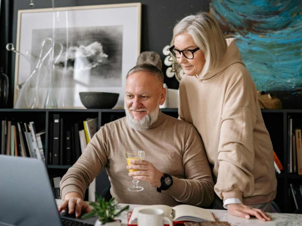 uomo seduto davanti al computer con un bicchiere in mano e donna in piedi alle sue spalle