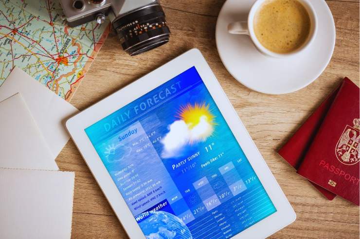 Tablet che mostra previsioni meteo poggiato su un tavolo accanto a passaporti, tazzina di caffe, macchina fotografica e mappa