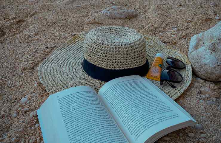 Un libro ed un cappello in spiaggia con crema solare ed occhiali da Sole