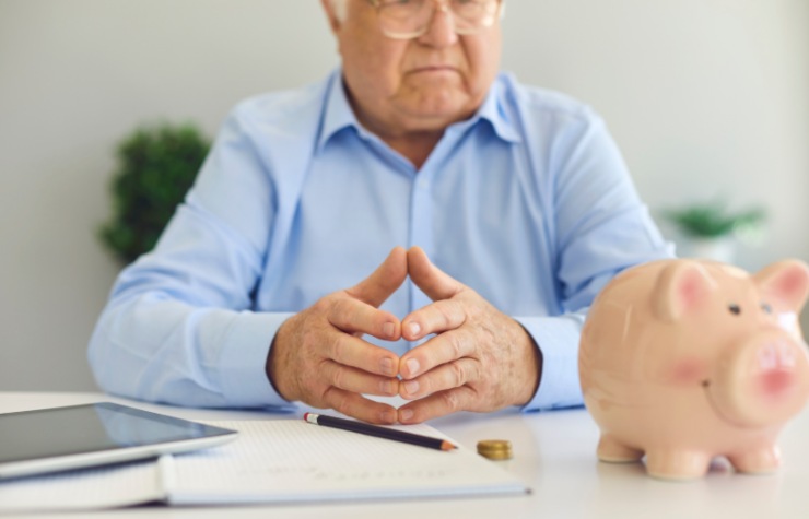 uomo anziano pensieroso con davanti un foglio, una matita e un salvadanaio