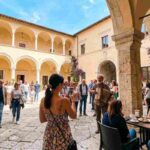 Turisti visitano un chiostro storico a L'Aquila in Abruzzo durante il ponte del 1° maggio, tra architettura medievale e atmosfera conviviale.