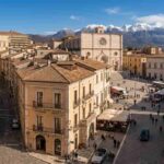 Veduta panoramica dall'alto del centro storico dell'Aquila, Abruzzo, con la Basilica di Collemaggio, Piazza Duomo e le montagne del Gran Sasso innevate sullo sfondo in una giornata di sole.