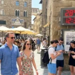Turisti a Firenze in Piazza della Signoria con un termometro digitale che segna 28°C il 25 Aprile, mostrando il caldo anomalo record.