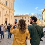 Turisti in centro storico a L’Aquila durante il ponte del 25 aprile 2026, con vista sulle vette innevate del Gran Sasso e cartelli informativi della Capitale Italiana della Cultura.