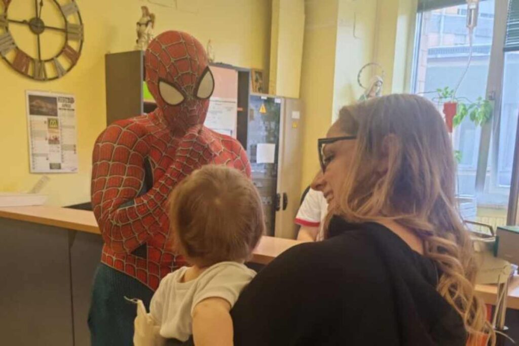 Un uomo vedtito da Spider-Man davanti ad un bambino tenuto in braccio da una donna sorridente, entrambi ritratti di spalle.