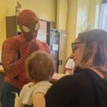 Un uomo vedtito da Spider-Man davanti ad un bambino tenuto in braccio da una donna sorridente, entrambi ritratti di spalle.