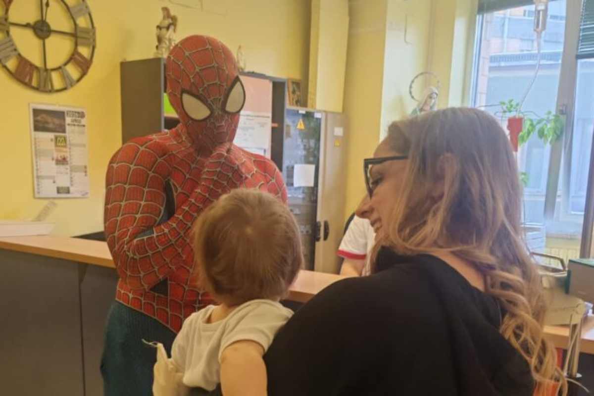Un uomo vedtito da Spider-Man davanti ad un bambino tenuto in braccio da una donna sorridente, entrambi ritratti di spalle.