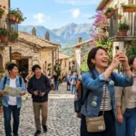 Turisti giapponesi sorridenti scattano foto in un borgo medievale dell'Abruzzo, montagne Gran Sasso sullo sfondo, luce solare estiva, turismo esperienziale 2026.