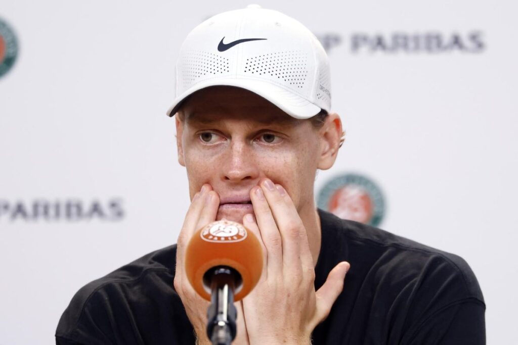 Jannik Sinner con una espressione stanca si tiene il volto con le mani davanti ad un microfono.
