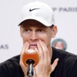 Jannik Sinner con una espressione stanca si tiene il volto con le mani davanti ad un microfono.