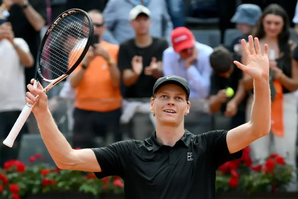 Jannik Sinner esulta con racchetta e braccia in alto con divisa nera.