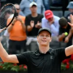 Jannik Sinner esulta con racchetta e braccia in alto con divisa nera.