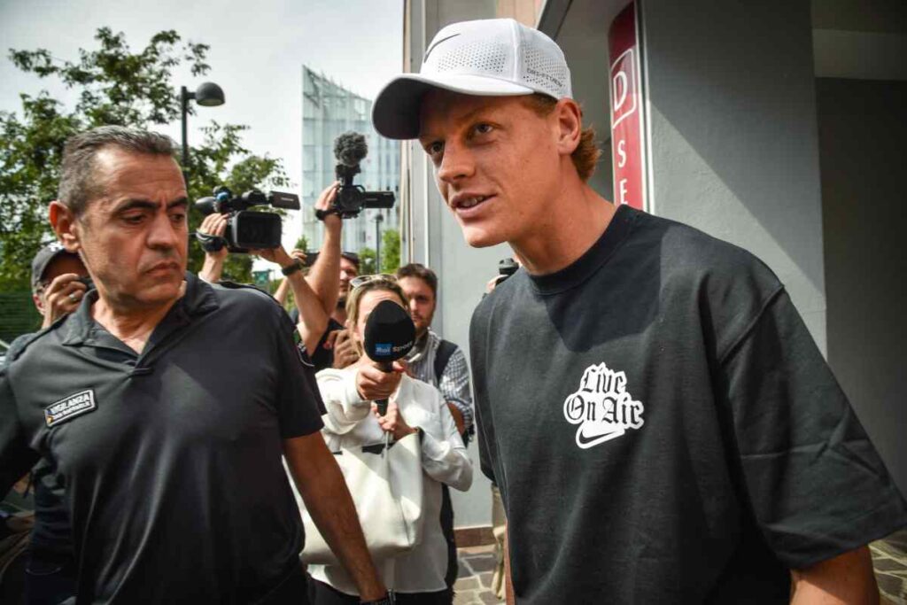 Jannik Sinner con t-shirt nera e berretto bianco al cospetto di giornalisti e fotografi in esterna.